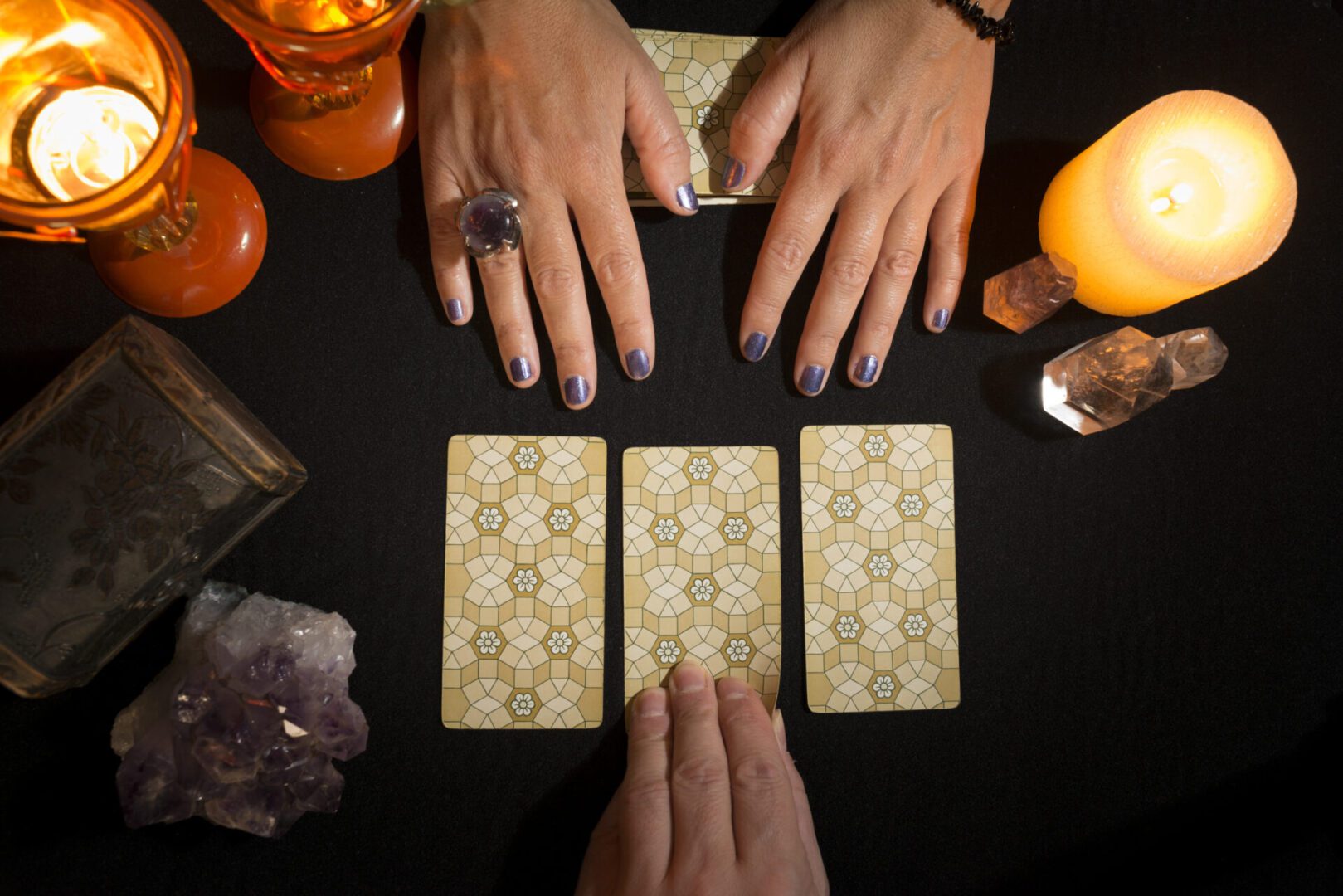 Detail of a woman's hands showing three tarot cards face down on a black card table, while another hand holds up one of the cards. Concept of divination session with tarot cards. View from above.